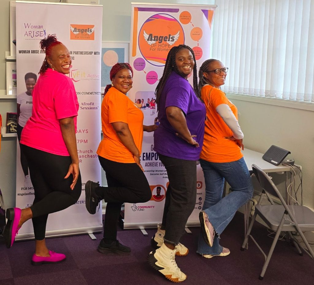 Four women pose smiling for the photo, kicking their front foot back. They wear bright pink, orange and purple t-shirts.